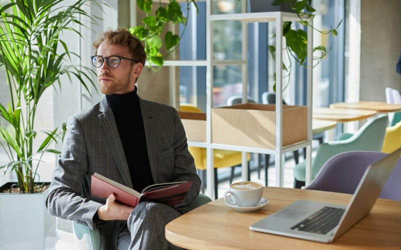 confident-young-business-coach-holds-a-book-and-looks-away-sitting-at-a-table-with-laptop-in-office.jpg